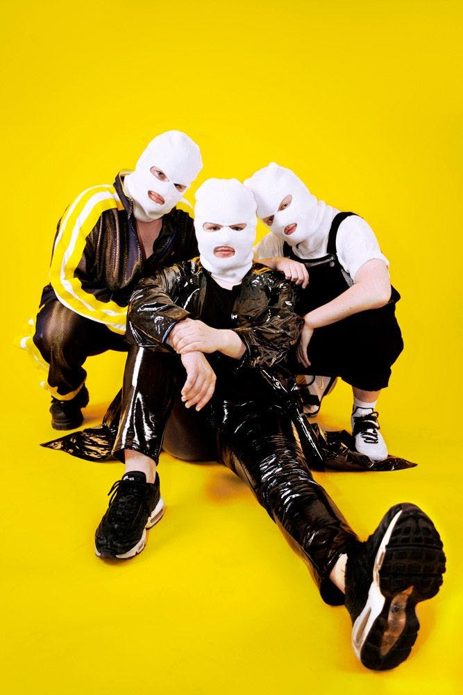 Three men on the ground that have ski masks on in front of a yellow backdrop